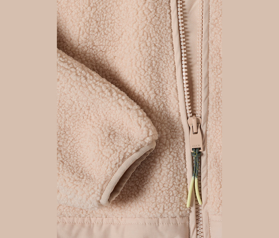Nahaufnahme einer beigen CECIL Teddy-Jacke, die flauschige Textur, einen Reißverschluss mit beigem Schieber und eine grün-gelbe Kordel neben einem gerippten Ärmelbündchen zeigt.