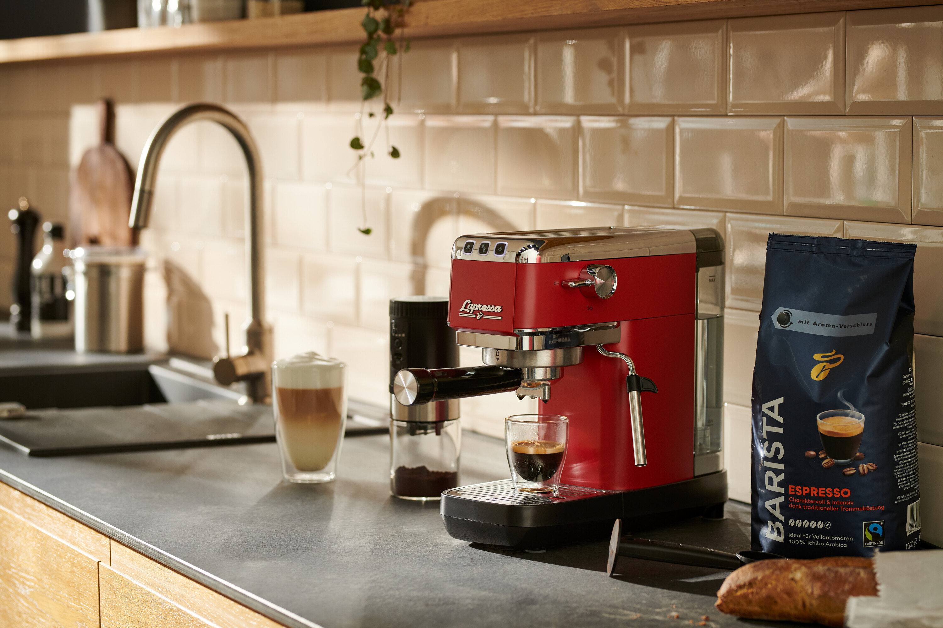 Eine rote »Lapressa« Siebträgermaschine bereitet Kaffee in einem Glas zu. Daneben stehen eine Kaffeemühle und eine Packung Kaffee.