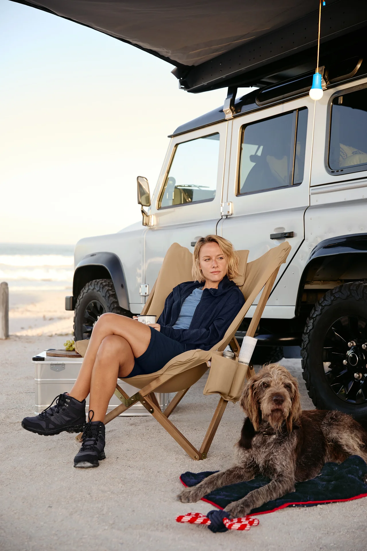 Eine Frau sitzt in einem Relaxsessel mit einem Becher. Neben ihr liegt ein Hund auf einer Hunde-Kuscheldecke mit einem Hunde-Spielzeug, Tau. Im Hintergrund sind ein Auto, eine LED-Camping-Lampe, eine 2-in-1-Funktionshose und eine Kapuzenjacke.