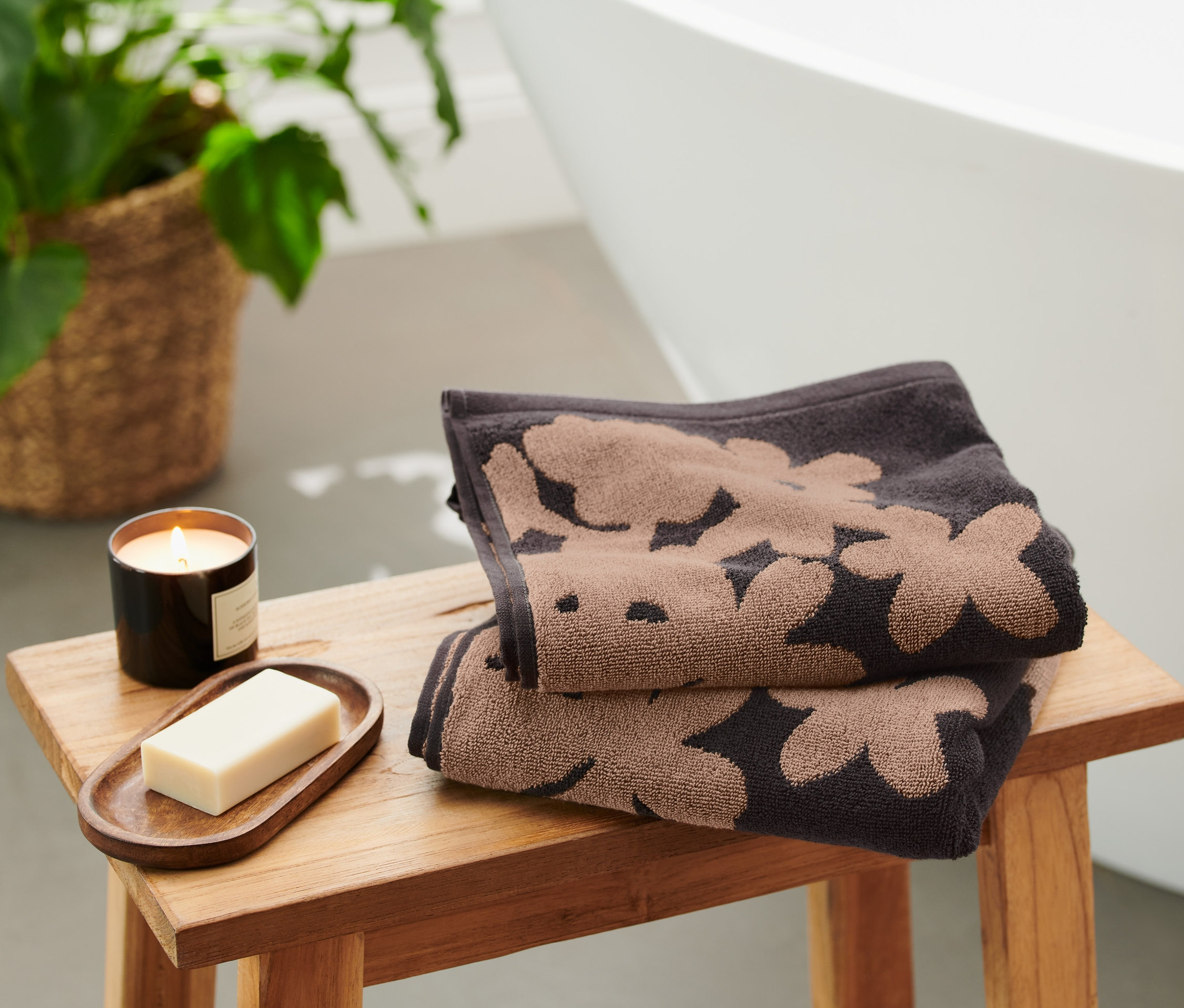 Zwei schwarze Jacquard-Handtücher mit Blumenmuster liegen gefaltet auf einem Holzhocker neben einer Kerze und Seife.