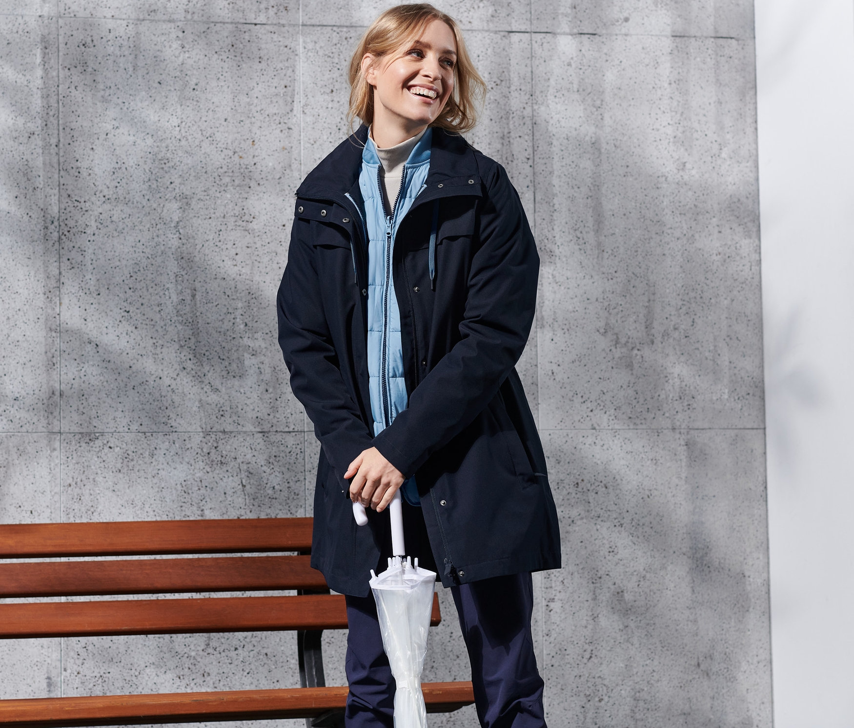 Eine lächelnde Frau steht mit einem Regenschirm und trägt einen blauen 3-in-1-Outdoormantel.