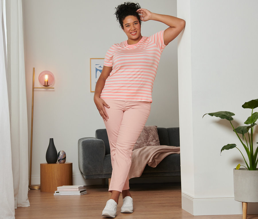 Frau posiert an eine Wand gelehnt in einem roséfarbenen, gestreiften T-Shirt, einer roséfarbenen Slimfit-Jeans und Veloursleder-Sneakers.