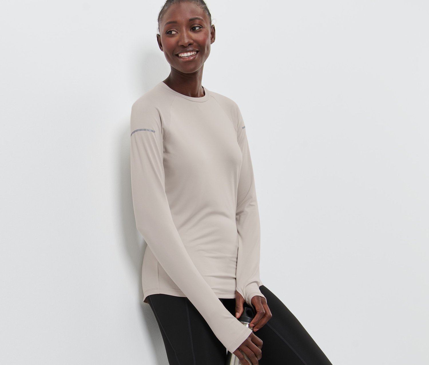 Frau lehnt an einer Wand und hält eine Metallwasserflasche. Sie trägt ein graues Langarm-Funktionsshirt und schwarze Leggings.