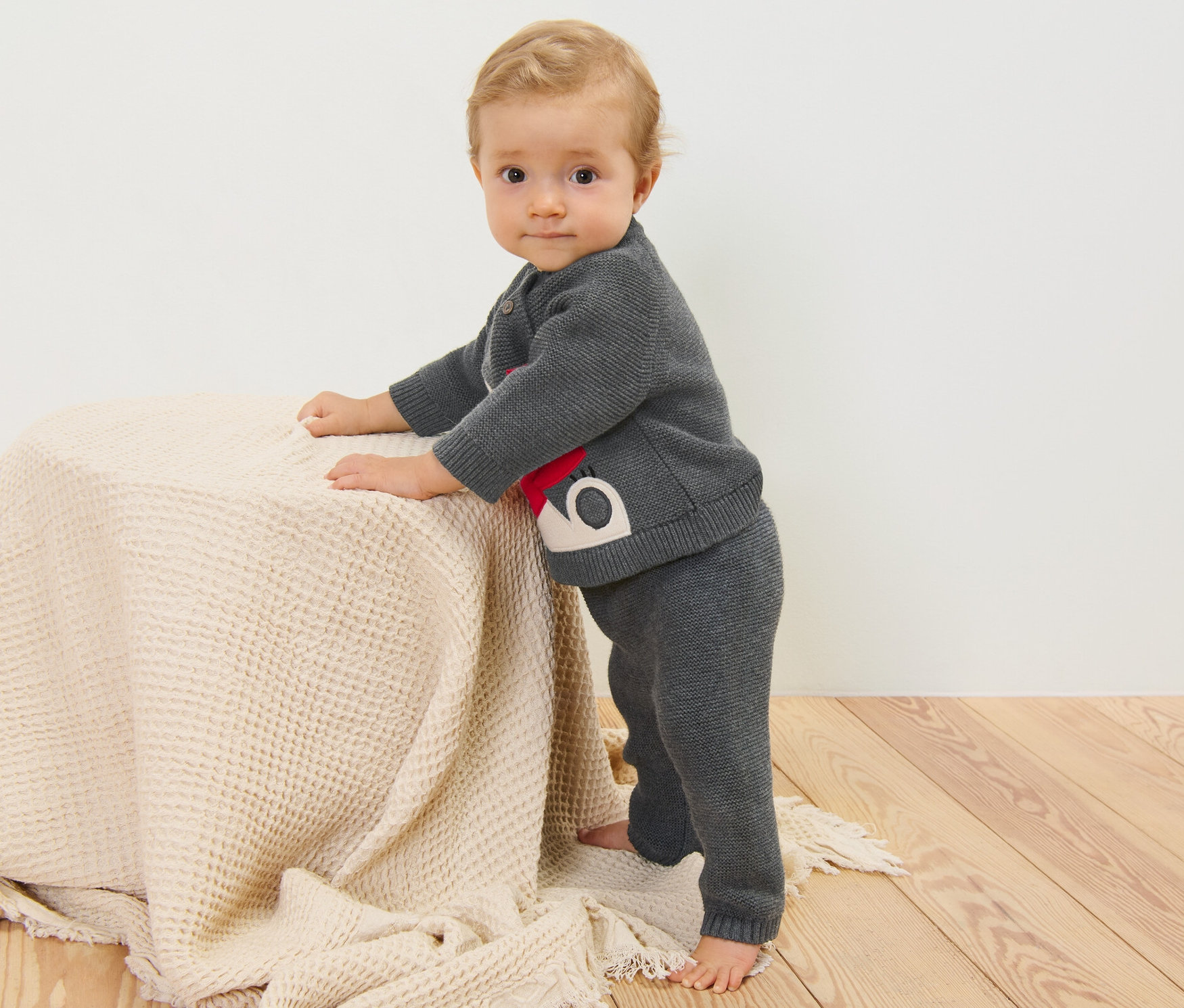 Baby steht und hält sich an einem Tisch fest. Es trägt eine Kinder-Strickhose.