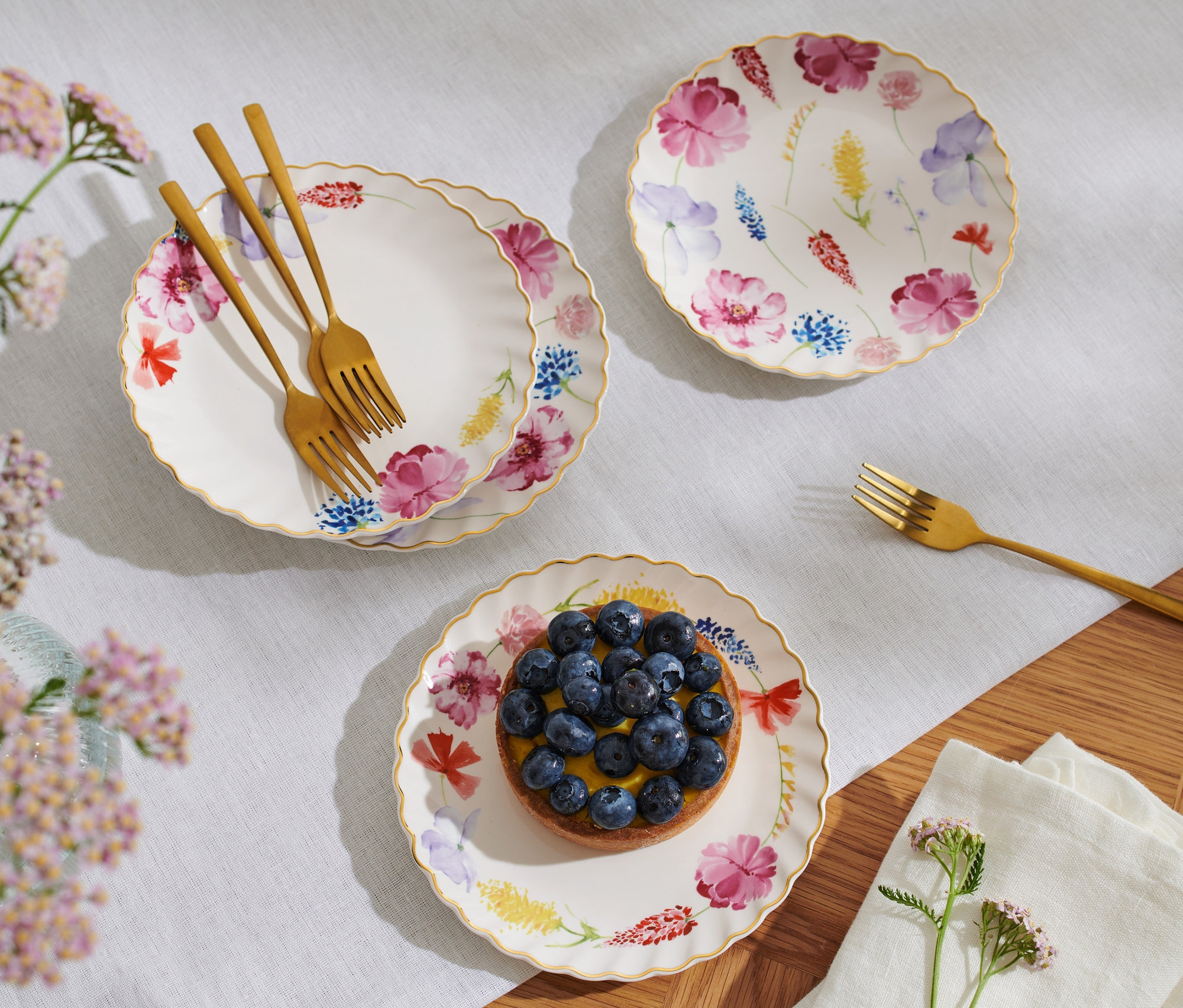 Drei Dessertteller mit Glanzgolddekor und Blumenmuster, einer mit Blaubeerkuchen, daneben liegen goldene Gabeln.