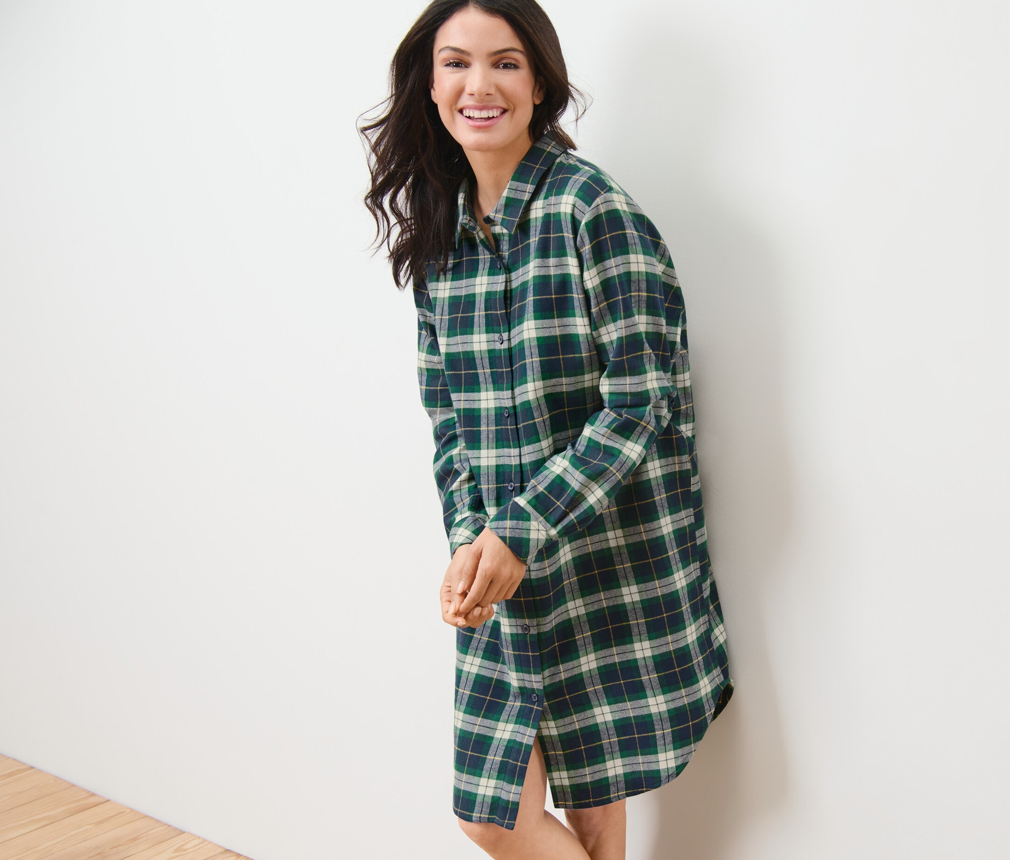 Lächelnde Frau, die sich an eine weiße Wand lehnt, trägt ein grünes Flanell-Nachthemd.