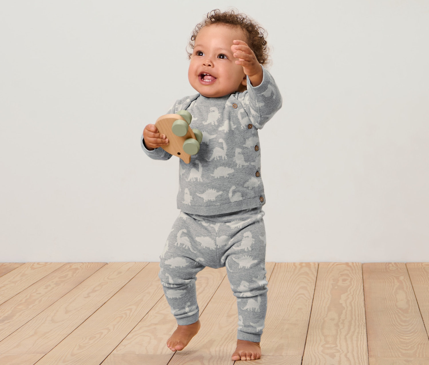 Ein Baby steht auf einem Holzboden und hält ein Holzspielzeug. Es trägt eine Baby-Strickkombi.