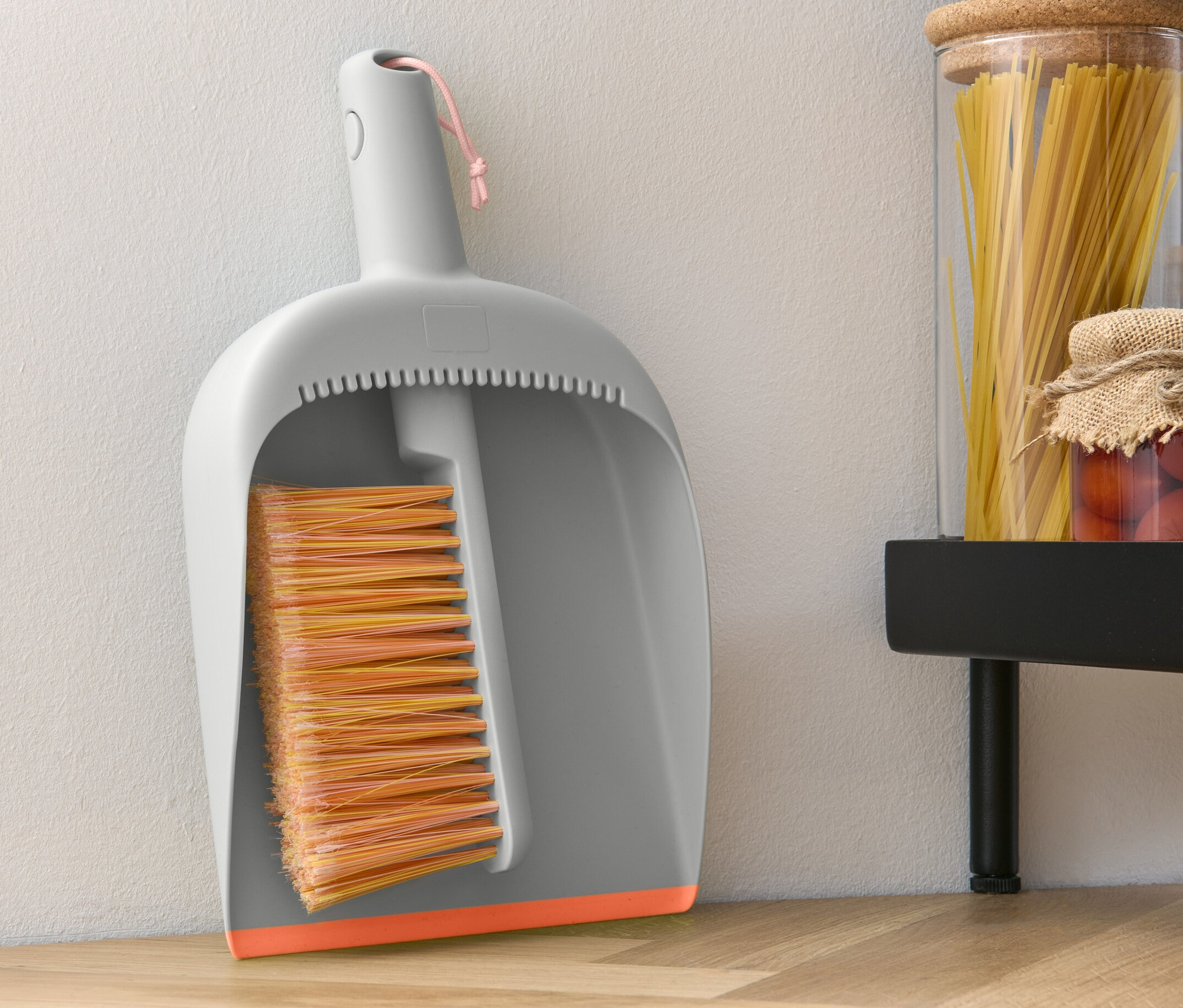 Grauer Handkehrer und Schaufel lehnen an einer Wand neben einem Regal mit Lebensmitteln.