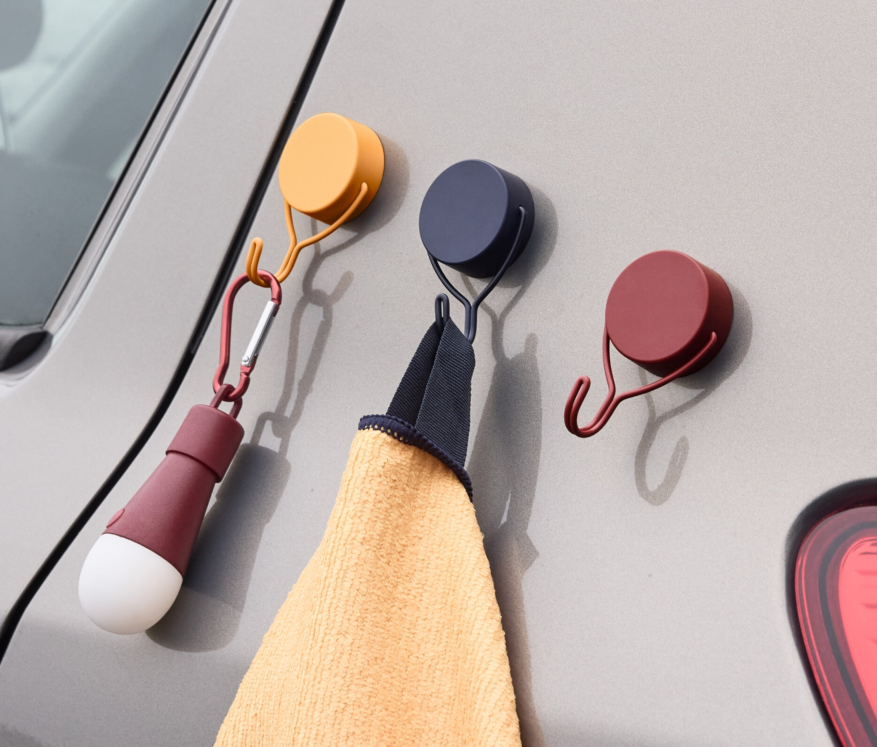 Drei Magnethaken sind an einem grauen Auto befestigt. An einem Haken hängt ein Handtuch, an dem anderen eine Lampe.