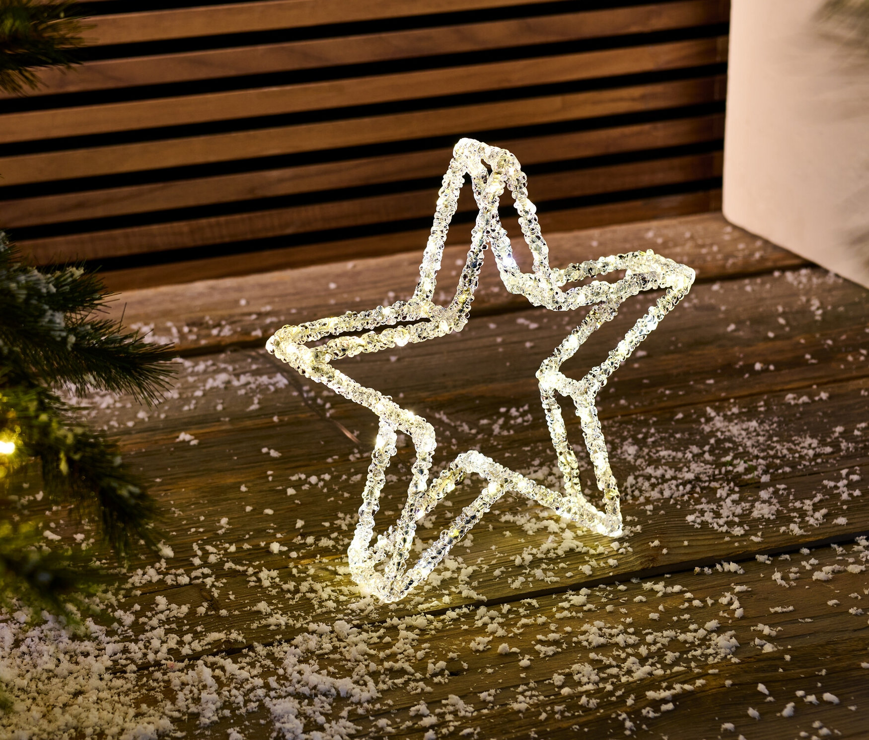 Ein LED-Dekostern steht auf einem mit Kunstschnee bedeckten Holzboden neben einem Weihnachtsbaum.