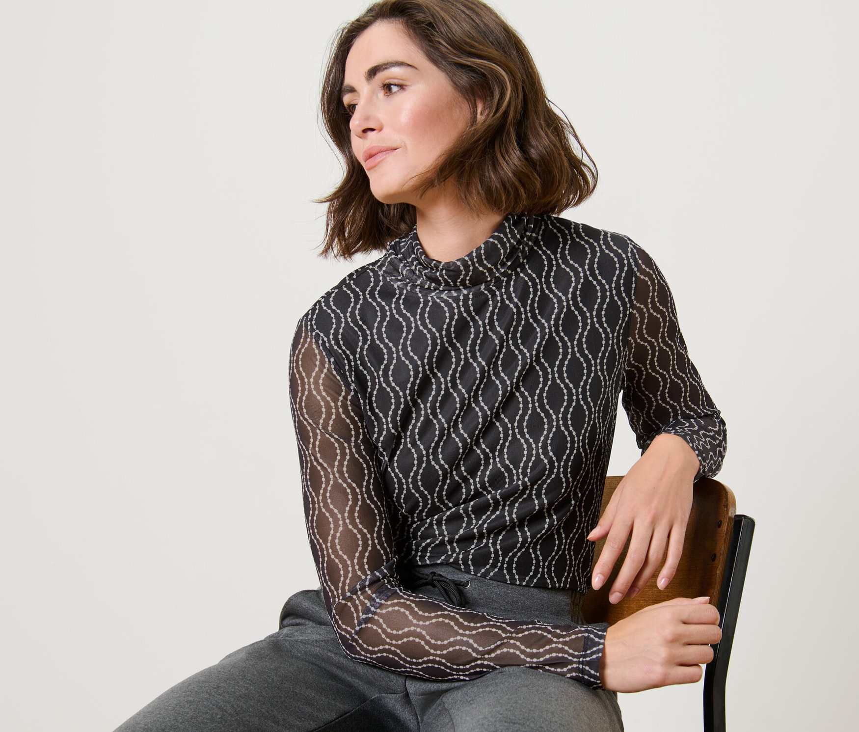 Frau sitzt auf einem Stuhl und trägt ein bedrucktes Mesh-Rollkragenshirt und graue Trainingshose.