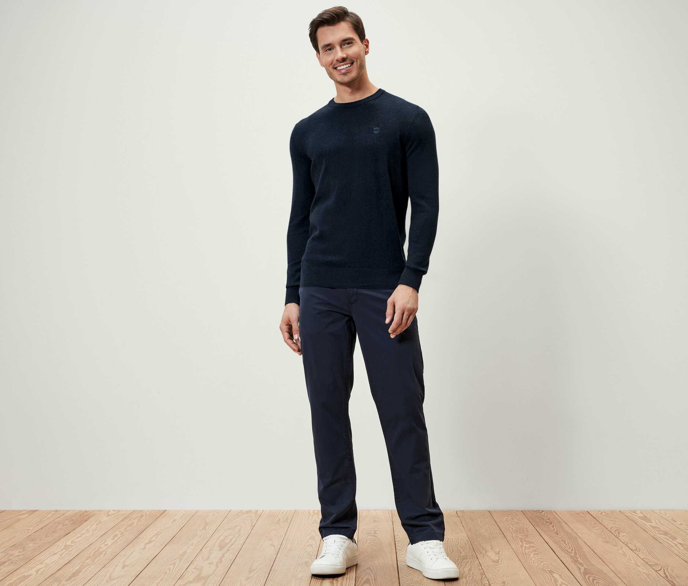 Ein Mann steht und trägt einen dunkelblauen Pullover mit Rundhalsausschnitt, dunkelblaue Hosen und weiße Turnschuhe.