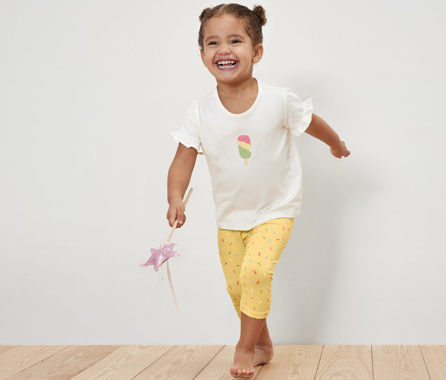 Lächelndes Mädchen posiert in einem T-Shirt mit einem Eis am Stiel und einer pink/gelben 2 ¾-Kinder-Leggings.