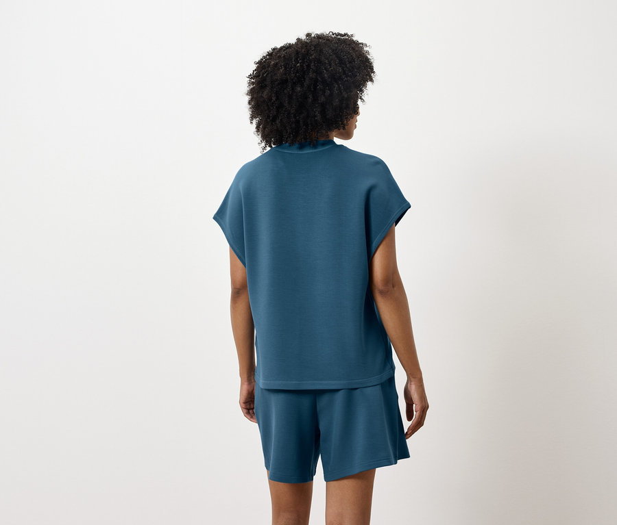 Eine Frau steht mit dem Rücken zur Kamera und trägt ein blaugrünes Oberteil und Shorts.