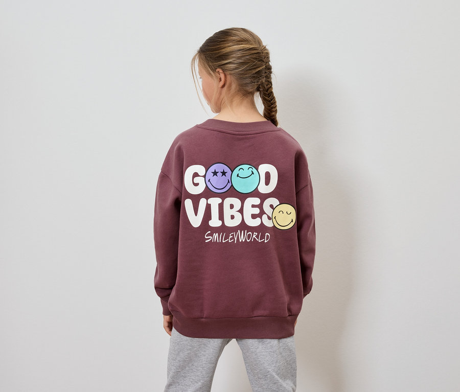 Mädchen mit Zopf trägt einen weinroten Pullover mit dem Aufdruck Good Vibes Smileyworld.