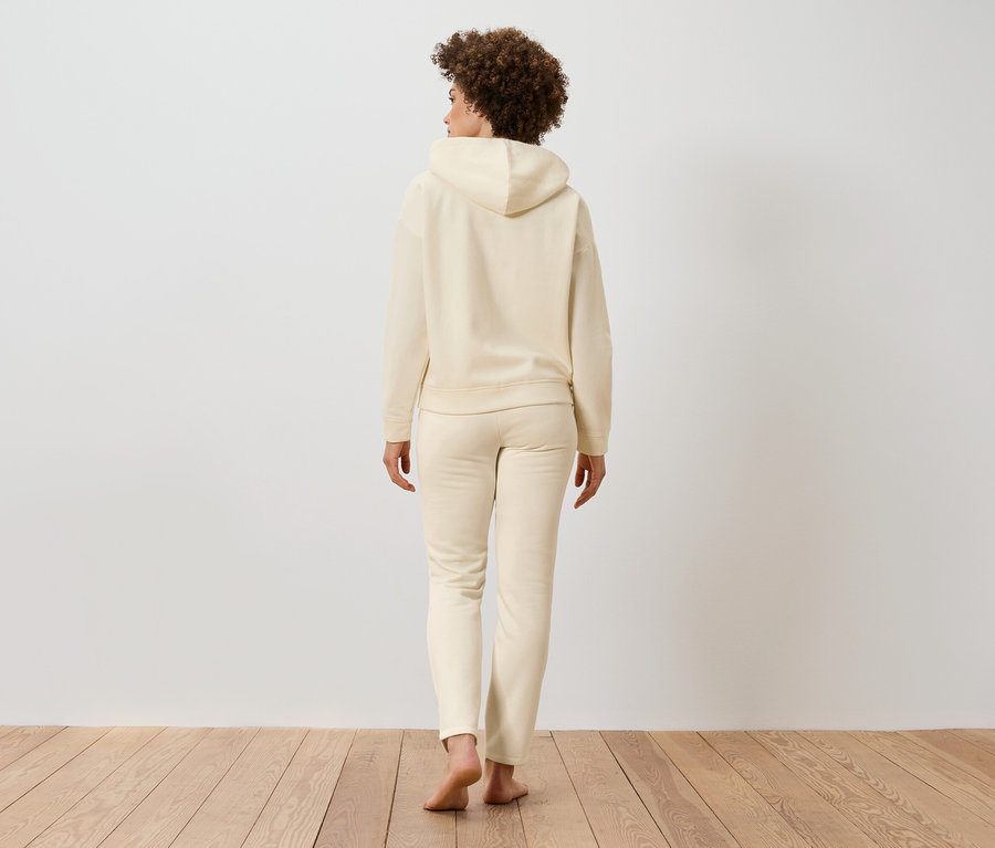 Frau mit lockigem Haar steht mit dem Rücken zum Betrachter, bekleidet mit einem beigen Kapuzenpullover und Jogginghose auf einem Holzboden.