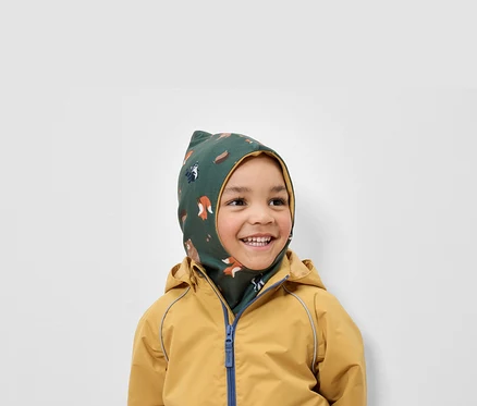 Lächelnder Junge trägt eine grüne Sturmhaube mit Tiermuster und eine gelbe Jacke.