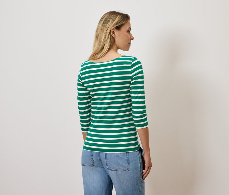 Eine Frau mit blonden Haaren steht mit dem Rücken zur Kamera und trägt ein grün-weiß gestreiftes Dreiviertelarmshirt und blaue Jeans.