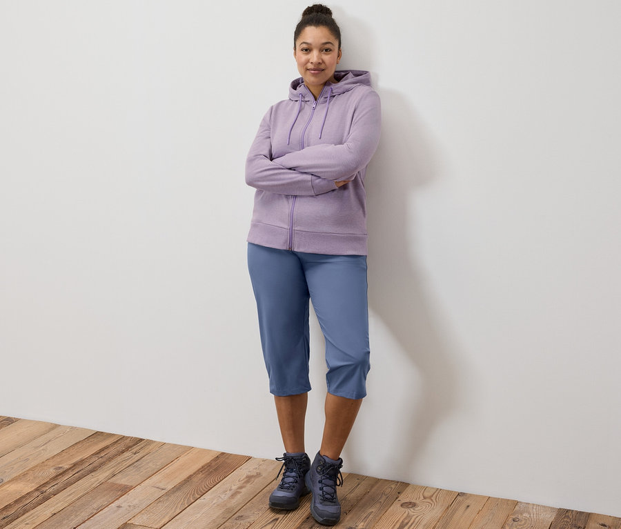 Eine Frau steht mit verschränkten Armen und trägt eine lila Sweatjacke und Funktionscapri.