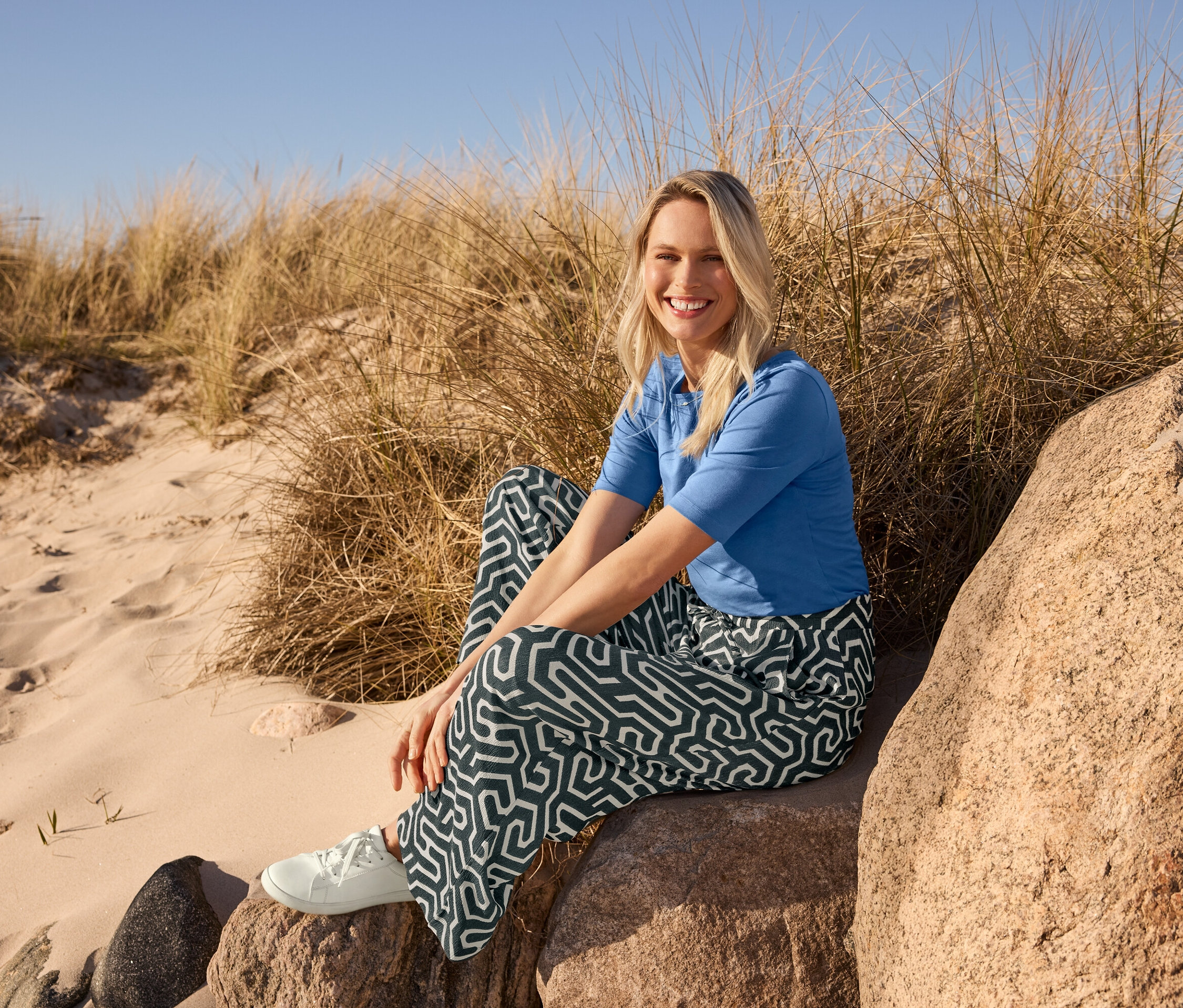 Eine Frau sitzt am Strand auf einem Stein und trägt ein blaues Halbarmshirt, eine bedruckte Schlupfhose in Crinkle-Optik und Ledersneaker.