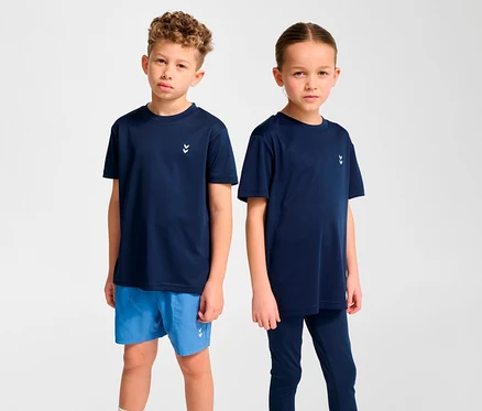 Zwei Kinder stehen nebeneinander. Der Junge links trägt ein dunkelblaues HUMMEL HMLJR Pulse Training-T-Shirt und blaue Shorts. Das Mädchen rechts trägt ein dunkelblaues T-Shirt und Leggings.
