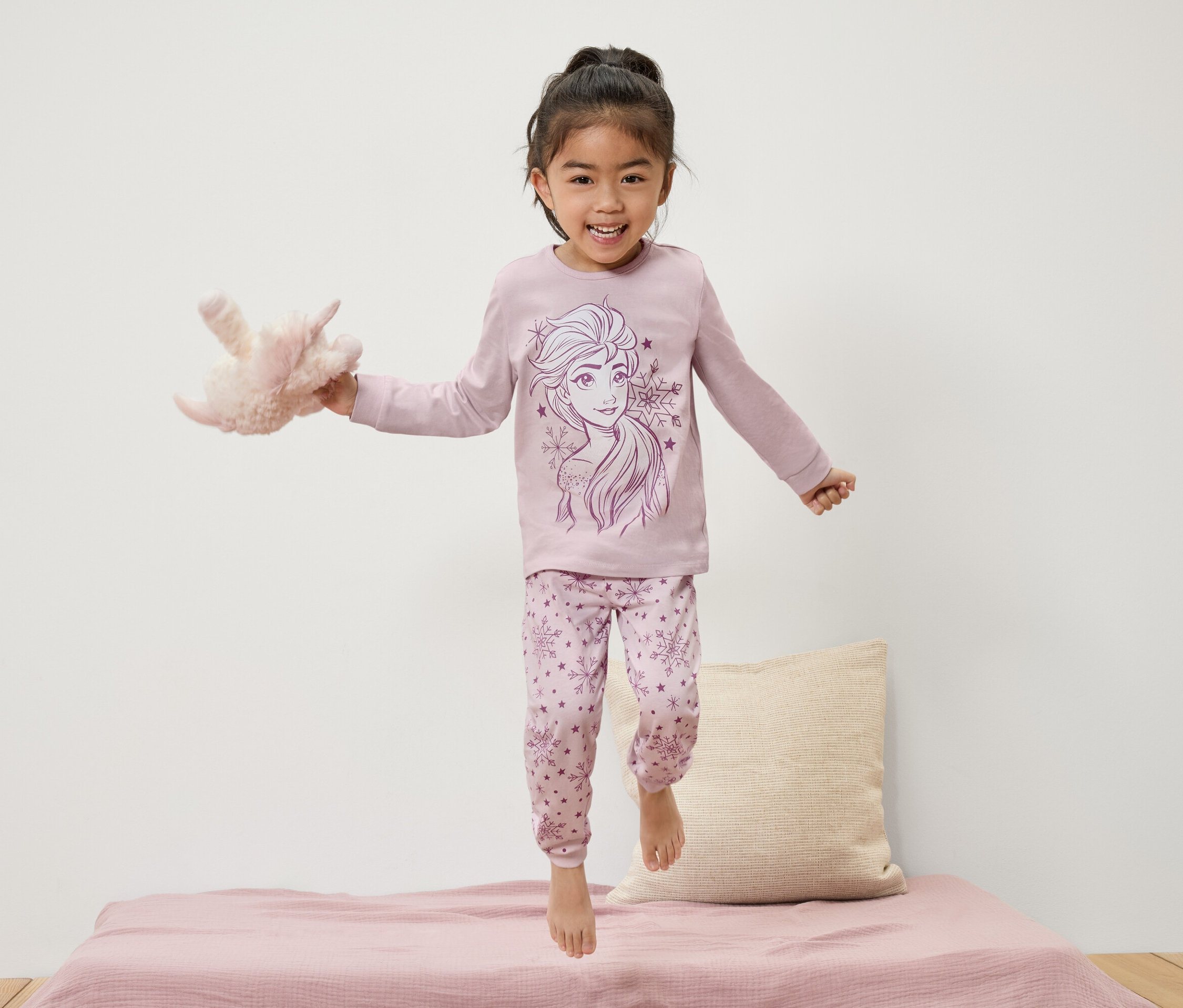 Mädchen im Elsa-Pyjama springt auf dem Bett und hält ein Plüschtier.