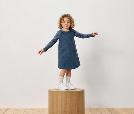 Ein Mädchen mit lockigem Haar steht auf einem Holzsockel. Sie trägt ein blaues Kleid und weiße Turnschuhe.