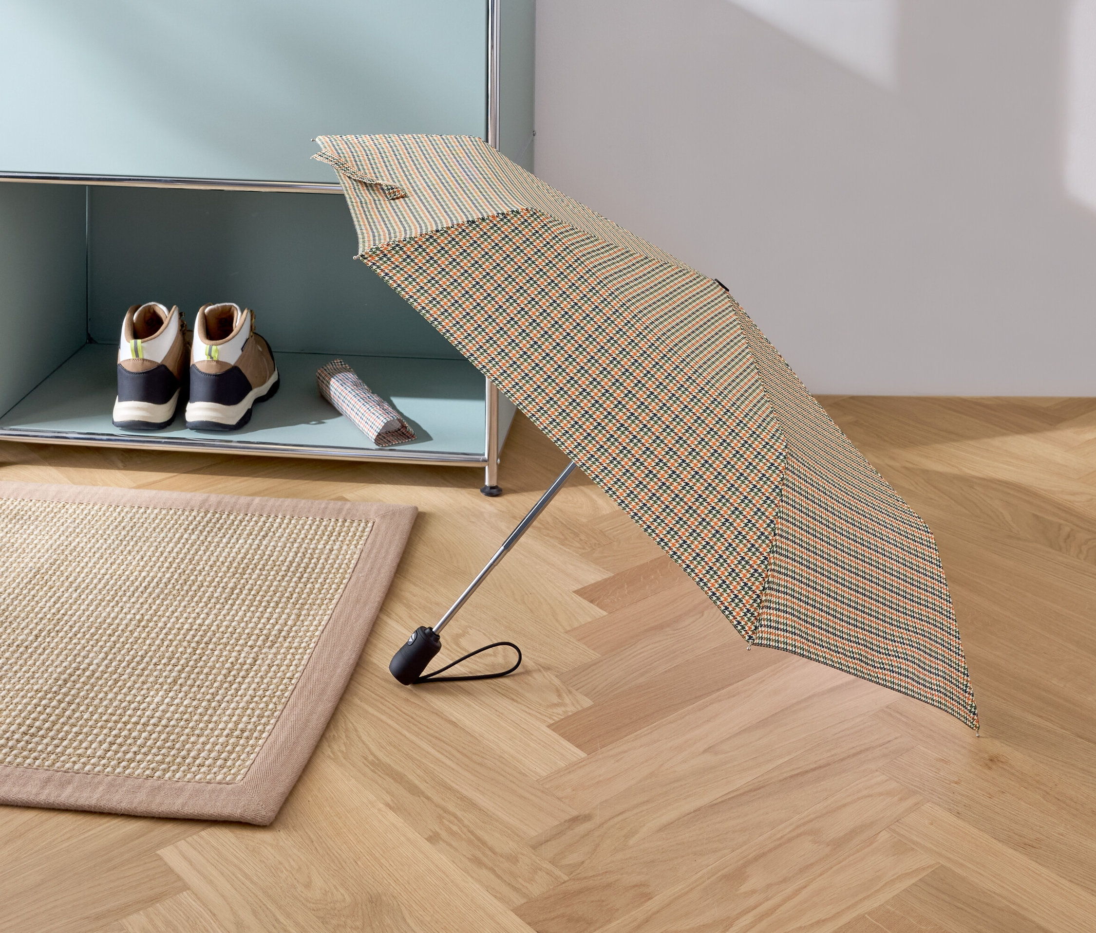 Ein geöffneter Regenschirm mit Karomuster steht auf einem Parkettboden. Im Hintergrund sind ein beiger Teppich, Schuhe und eine Tasche in einem Regal zu sehen.