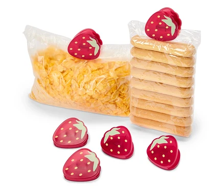 Eine Tüte Cornflakes und eine Packung Kekse sind mit Erdbeer-Clips verschlossen. Fünf weitere Erdbeer-Clips liegen im Vordergrund.