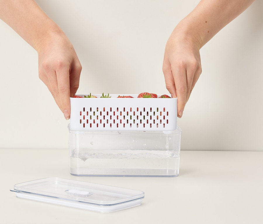 Hände platzieren einen weißen Behälter mit Erdbeeren auf einem durchsichtigen Behälter mit Wasser.