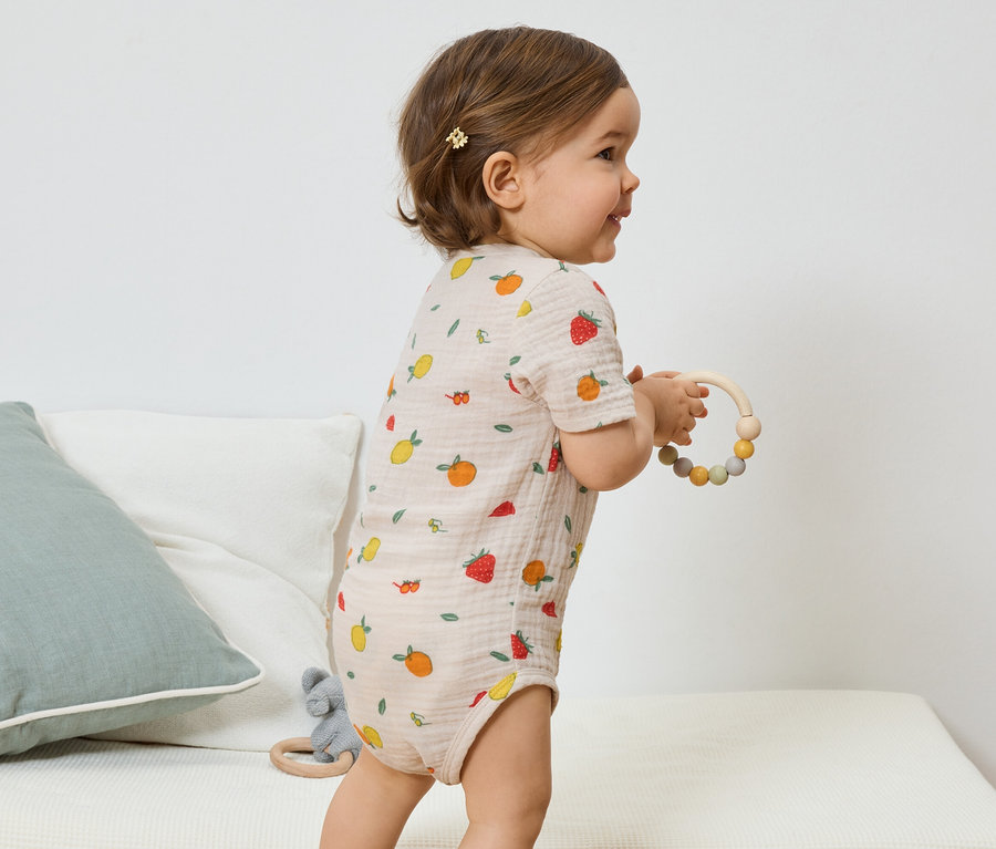 Eine Seitenansicht eines Babys mit einer hellbraunen Haarspange, das einen hellbraunen, kurzärmeligen Body mit Fruchtmuster trägt und einen hölzernen Beißring hält.