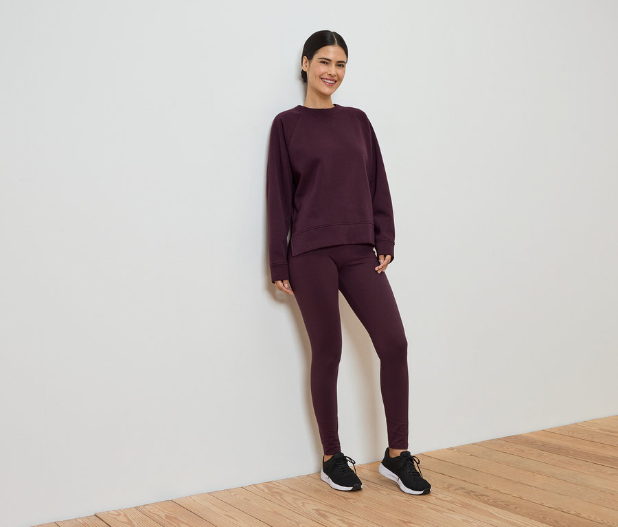 Frau in weinrotem Trainingsanzug, bestehend aus Leggings und Pullover, lehnt an einer weißen Wand.