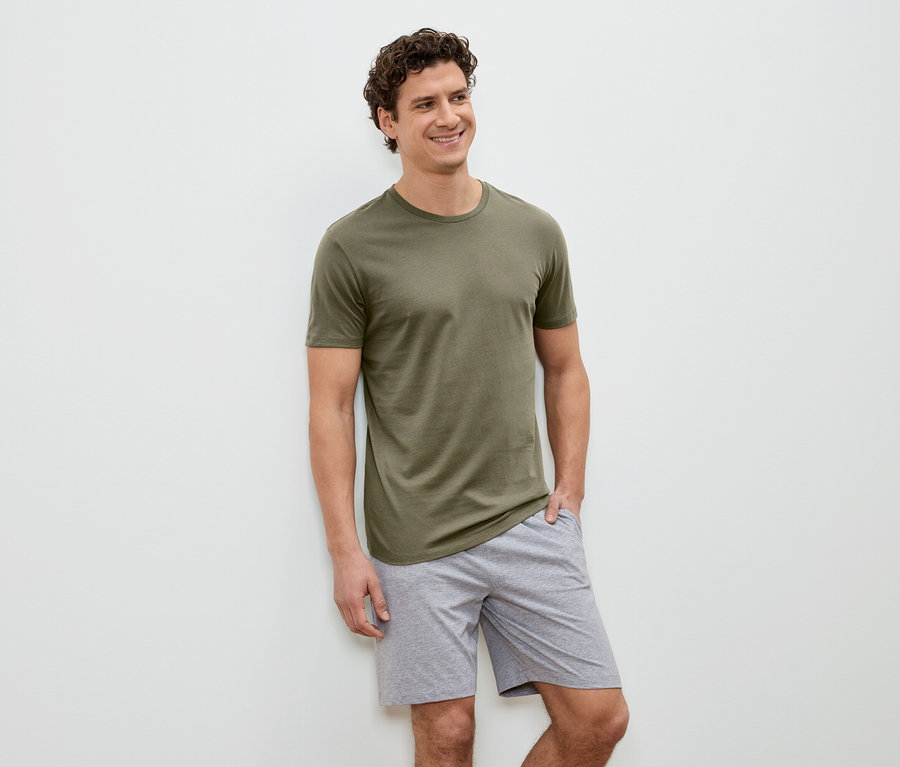 Mann mit lockigem Haar, olivgrünem T-Shirt und grauen Shorts steht vor einer weißen Wand und lächelt.