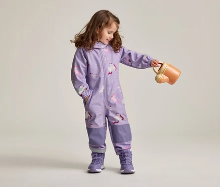 Mädchen mit braunen, lockigen Haaren in lila Overall mit Einhornmuster und lila Schuhen hält eine orangefarbene Gießkanne.