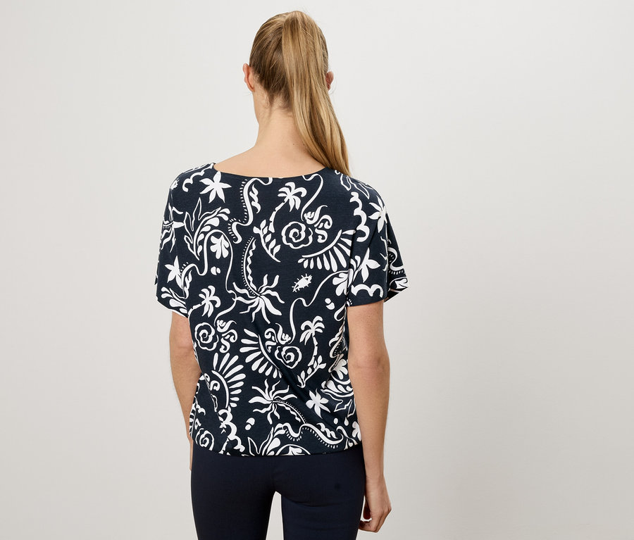 Rückansicht einer Frau mit blondem Pferdeschwanz, die ein marineblaues T-Shirt mit weißem Blumenmuster und marineblaue Hosen trägt.