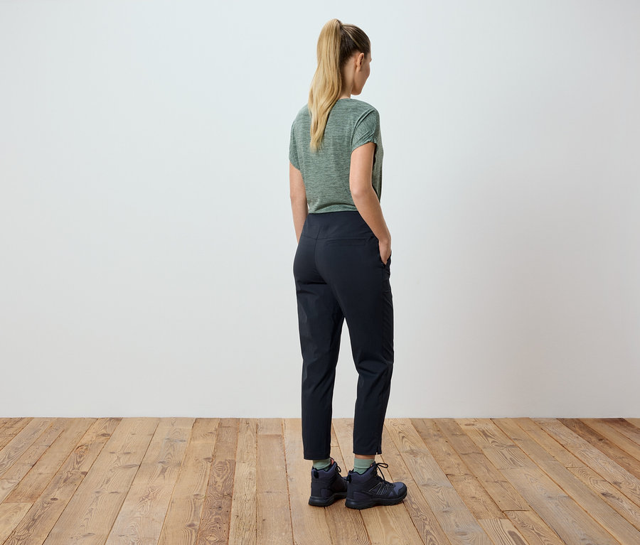 Frau mit blonden Haaren trägt ein grünes T-Shirt und schwarze Hose, die Hände in den Taschen.