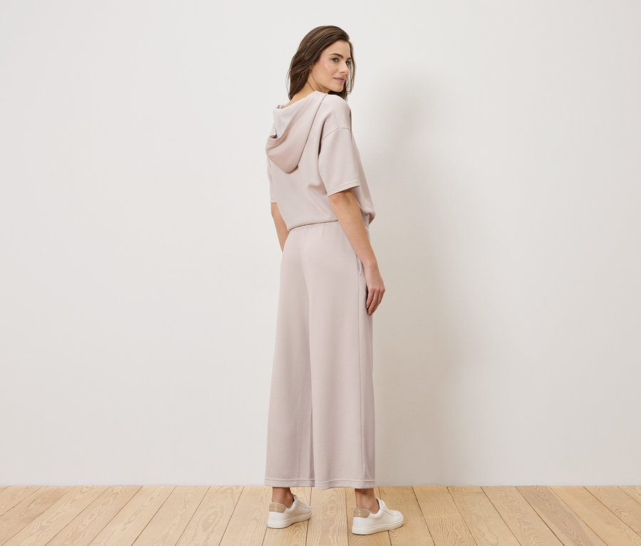 Frau steht in einem hellrosa Hosenanzug und weißen Turnschuhen, den Kopf über die Schulter gedreht.