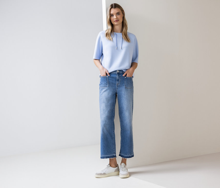 Eine Frau steht mit ihren Händen in den Taschen vor einer weißen Wand. Sie trägt ein hellblaues Kurzarmshirt, eine blaue Jeans und weiße Turnschuhe.