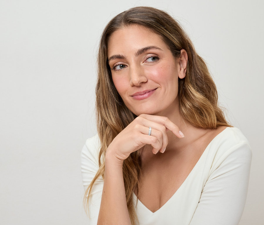 Frau mit langem, gewelltem Haar und weißem Oberteil, die einen silbernen Ring am Finger trägt und nachdenklich zur Seite blickt.