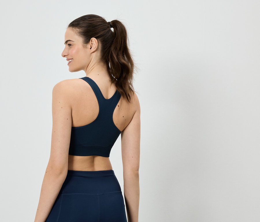 Frau mit Pferdeschwanz, von hinten zu sehen, trägt einen marineblauen Sport-BH und Leggings vor einem weißen Hintergrund.