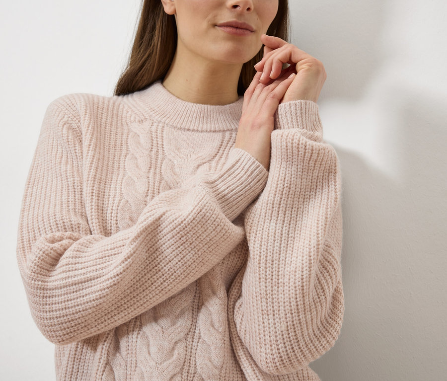 Frau mit den Händen am Gesicht trägt einen hellrosa Zopfstrickpullover vor einer weißen Wand.