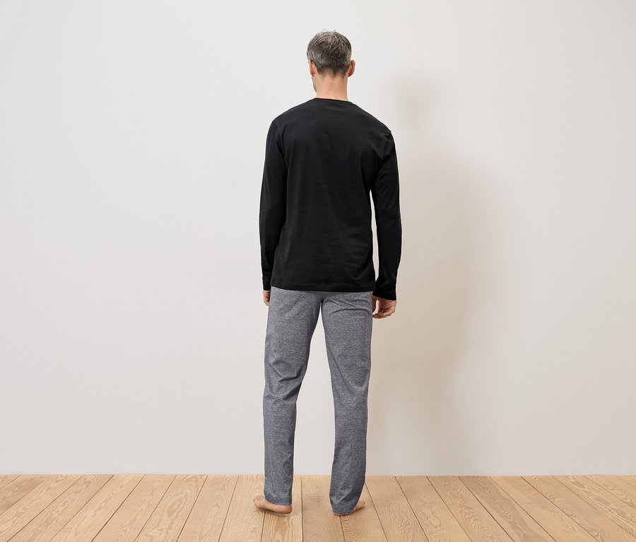 Mann von hinten in schwarzem Langarmshirt und grauer Hose vor einer Wand auf Holzboden.