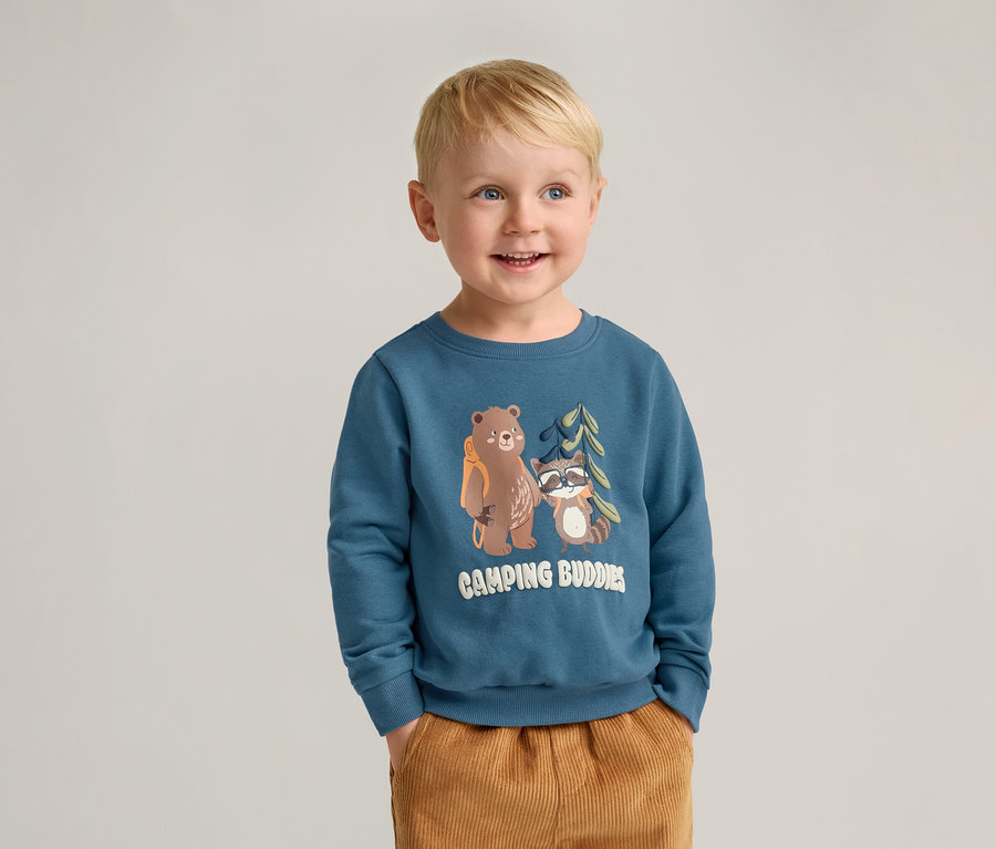 Lächelnder Junge mit blonden Haaren trägt ein blaues Sweatshirt mit Bär- und Waschbär-Motiv und braune Hosen.