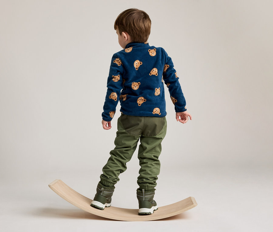 Ein kleiner Junge, von hinten gesehen, steht auf einem hölzernen Balance Board. Er trägt ein dunkelblaues Langarmshirt mit Bärengesichtern, olivgrüne Hosen und dunkelgrüne Stiefel.