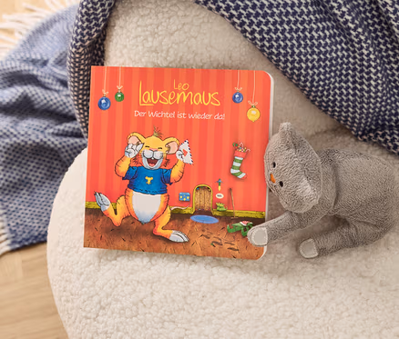 Auf einem weißen Hocker liegen ein Bilderbuch mit einem Cartoon-Löwen und ein graues Stofftier in Katzenform.