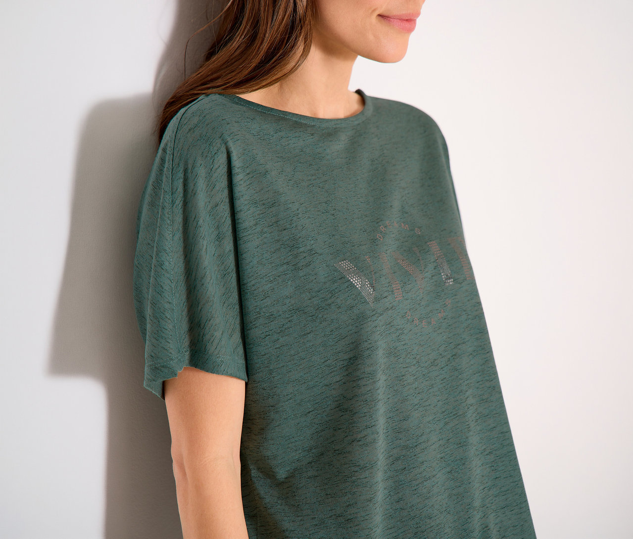 Frau lehnt an einer Wand und trägt ein grünes T-Shirt mit der Aufschrift VIVID.
