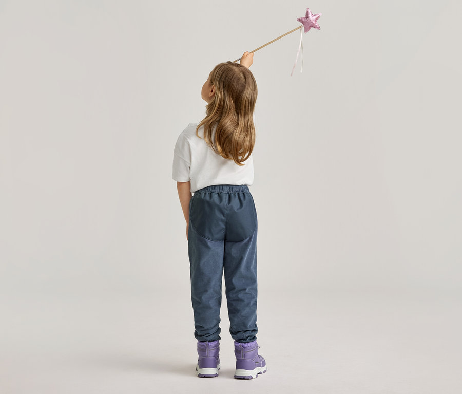Rückansicht eines Mädchens mit blonden Haaren, das einen Zauberstab mit rosa Stern hält. Sie trägt blaue Hosen und lila Stiefel.