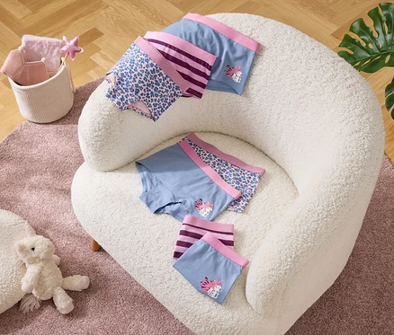Auf einem Sessel liegen Unterhosen in Blau mit lila Bündchen und Mustern. Daneben ein Korb mit Textilien und ein Teddy auf einem Teppich.