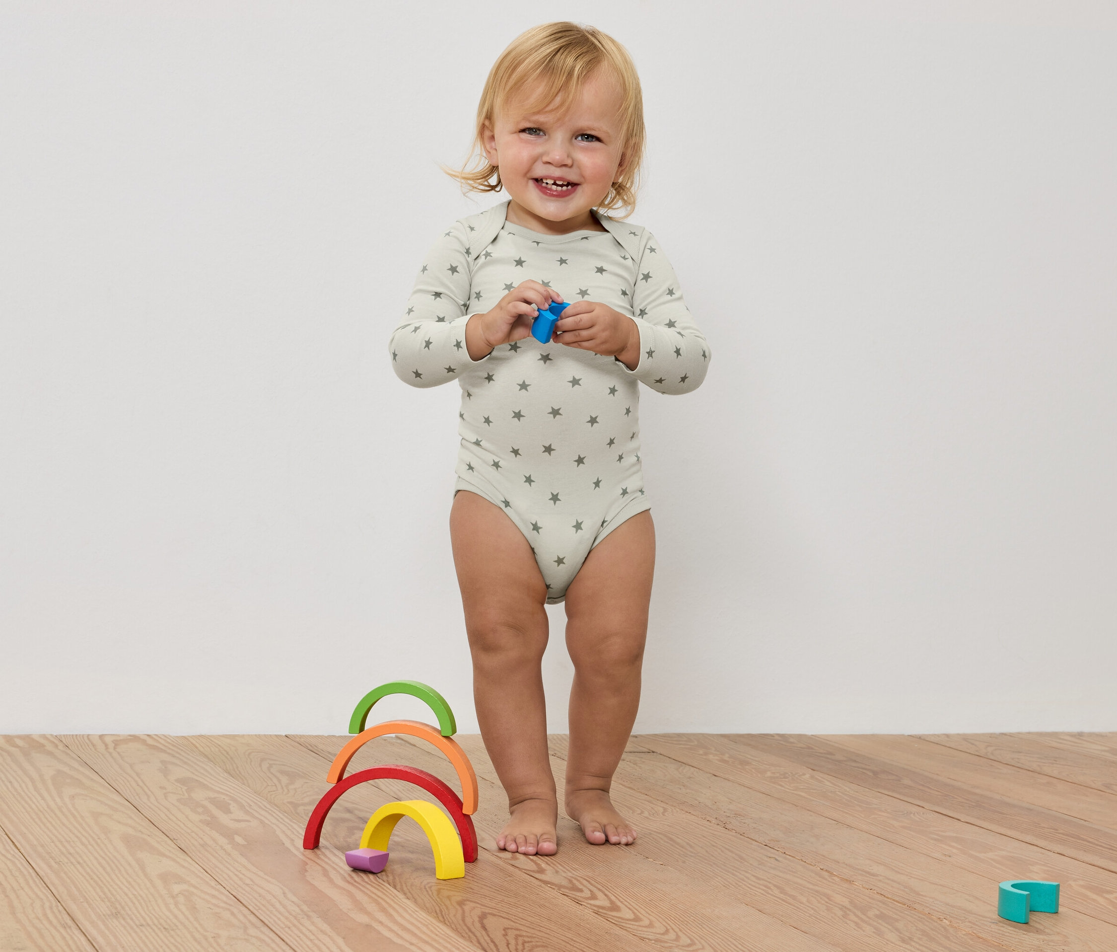 Ein lachendes Kleinkind im Sternenbody hält ein Spielzeug in den Händen, vor einem bunten Holzregenbogen auf einem Holzboden.