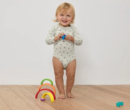 Ein lachendes Kleinkind im Sternenbody hält ein Spielzeug in den Händen, vor einem bunten Holzregenbogen auf einem Holzboden.